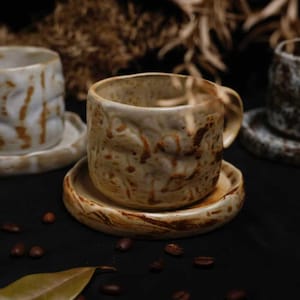 以下が含まれることがあります： 手作りのセラミックコーヒーカップとソーサーのセット。カップとソーサーは、ベージュと茶色のカラーパレットのテクスチャード加工された有機的なデザインです。コーヒー豆と葉が散らばっています。
