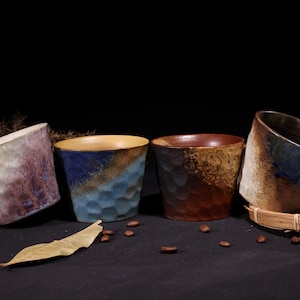 以下が含まれることがあります： ユニークなテクスチャの表面と色のグラデーションを持つ、手作りのセラミックマグカップ4つ。マグカップは、青、茶色、紫など、さまざまな色があります。1つのマグカップには織り込まれたハンドルが付いています。コーヒー豆と葉がマグカップの周りに散らばっています。