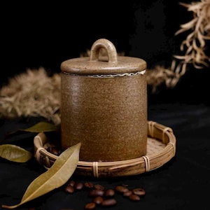 May include: A brown ceramic jar with a lid and handle, sitting on a small woven tray. The jar is cylindrical and has a speckled texture. Coffee beans and dried leaves are scattered around the base, against a dark background.