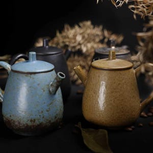 以下が含まれることがあります： さまざまな色の陶器のティーポットのコレクション。手前には、茶色のベースの青いティーポットと、茶色のティーポットがあります。他のティーポットが背景に見えます。ティーポットは、ハンドルと注ぎ口を備えたクラシックなデザインです。