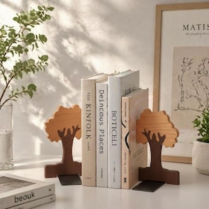 Puede incluir: Un par de sujetalibros de madera con forma de árbol sostienen libros. Los sujetalibros son de color marrón claro con una base marrón oscuro. Los libros tienen cubiertas blancas y beige con títulos como "The Kinfolk".