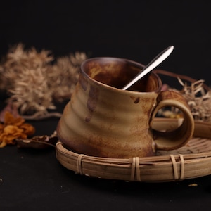 May include: A ceramic mug with a brown and tan swirl design, resting on a small woven tray. A silver spoon is inside the mug. The mug has a large handle. Dried plants and leaves are in the background.