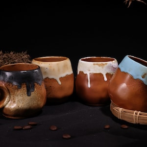 Puede incluir: Cuatro tazas de cerámica hechas a mano con diseños únicos. Las tazas presentan una base marrón con colores contrastantes en el borde, incluyendo negro, blanco y azul. Cada taza tiene un asa grande y curvada. Las tazas están dispuestas sobre una superficie oscura con granos de café.