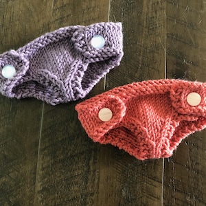 Könnte beinhalten: Zwei Paar gestrickte Baby-Windelüberzüge. Einer ist lila und der andere orange. Beide haben weiße Druckknöpfe.