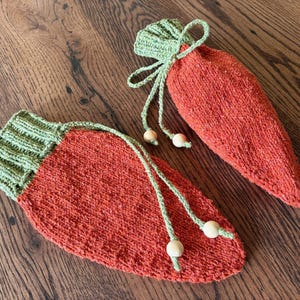 May include: Two carrot-shaped knitted pouches. The main body is orange, with a green knitted top and drawstring. The drawstring is green with wooden beads at the ends. One pouch is open, and the other is closed.