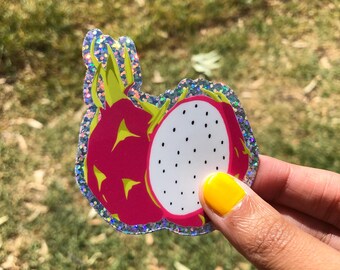 Könnte beinhalten: Ein glitzernder Aufkleber mit einer leuchtend rosa Drachenfrucht, mit einem weißen Inneren, das mit schwarzen Samen übersät ist. Der Aufkleber hat einen silbernen Glitzerrahmen. Der Hintergrund ist verschwommenes grünes Gras. Eine Hand hält den Aufkleber.