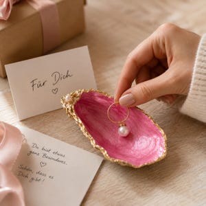 Könnte beinhalten: Eine dekorative Austernschale als Schmuckschale mit leuchtend pinkfarbenem Inneren und goldfarbenen Rändern, verziert mit einer kleinen Perle. Ein goldener Creolen-Ohrring mit Perlenanhänger wird darüber gehalten, neben Karten mit deutschem Text.