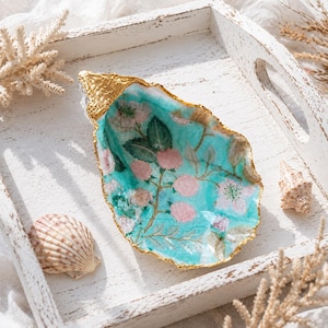 Könnte beinhalten: Dekorative Austernschale mit vergoldetem Rand. Das Innere zeigt einen türkisfarbenen Hintergrund mit einem Blumenmuster in Rosa und Weiß. Die Schale liegt auf einem weißen Holztablett mit Muscheln.