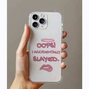 May include: Clear phone case with a pink halo, the words "OOPS! I ACCIDENTALLY SLAYED." and a pink lip graphic. The case is held by a hand against a neutral background. The phone case is designed to protect the phone.