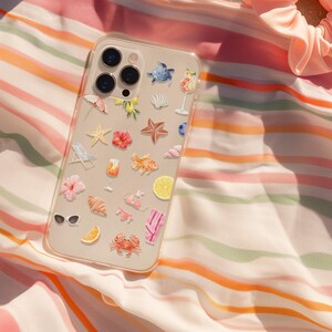 May include: A clear phone case adorned with beach-themed stickers, including starfish, flowers, and a crab, rests on a striped beach towel. A peach-colored scrunchie sits nearby on the sand, creating a summery scene.