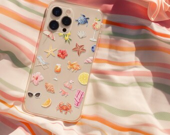 May include: A clear phone case adorned with beach-themed stickers, including starfish, flowers, and a crab, rests on a striped beach towel. A peach-colored scrunchie sits nearby on the sand, creating a summery scene.