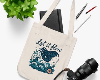 May include: Cream-colored tote bag with the text "Let it flow" and a graphic of a woman's silhouette with flowing dark blue hair, waves, and a flower. A black notebook, pens, and a camera are also visible.