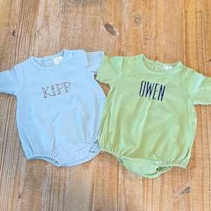 May include: A light blue short-sleeve romper with the name KIPP embroidered in red and a light green short-sleeve romper with the name OWEN embroidered in dark blue. Both garments feature a round neckline and snap closures at the bottom.