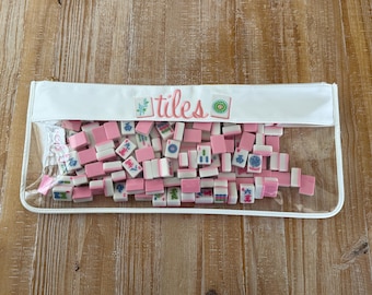 May include: A clear zippered pouch filled with pink and white Mahjong tiles. The pouch has a white top with the word "tiles" in pink script. The tiles are scattered inside the pouch, ready for play.
