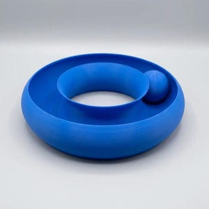 May include: A bright blue circular toy featuring a smooth, hollow ring structure with a central opening and a single, matching blue sphere resting within the curved track. The object has a matte finish and a continuous, rounded, donut-like shape.