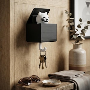 May include: A black wall-mounted storage box featuring a white cat figure that pops up when a set of keys is hung on the white hook below. The cat has black painted eyes, whiskers, and a smiling mouth. The text PAW-FECT HOOKS Your Keys, Safely Home is visible.