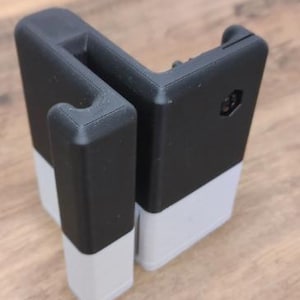 May include: A black and light gray L-shaped plastic bracket with a hook-like extension on one side. The object features a smooth, matte finish with visible 3D printed layer lines, a hexagonal cutout on the side, and a sturdy, angular construction.