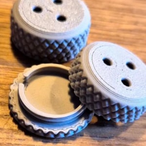 May include: Three light gray circular objects with a textured diamond pattern on the sides. Two of the objects are closed with three small holes on the top surface, while one is open, revealing a flat interior base and a circular rim structure.
