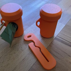 May include: Orange pet waste bag dispensers with a matching orange bag clip. One dispenser has a green bag partially pulled out. The dispensers have a cylindrical shape with a rounded top and a small handle.