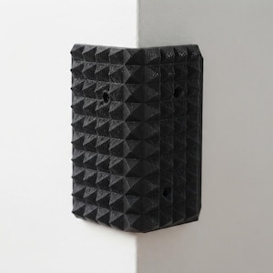 May include: A black L-shaped corner guard featuring a textured surface with a repeating pattern of small raised pyramids. The guard has pre-drilled circular holes for mounting and is positioned flush against the corner of a white wall.