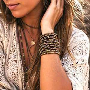 May include: A multi-strand beaded bracelet in shades of brown, green, and gold. The bracelet is wrapped around the wrist, showcasing the intricate details of the small beads. Several necklaces with coin pendants are also visible.