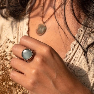 May include: A round, light blue gemstone ring with a bronze-colored band. The ring is worn on a hand, with a matching pendant necklace visible in the background. The jewelry has a vintage or bohemian aesthetic.