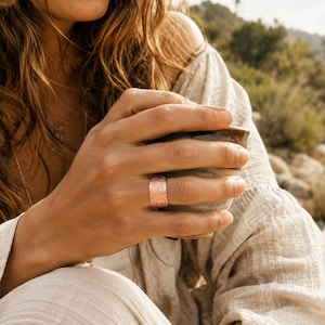 May include: A wide, rose gold-toned ring featuring a textured, hammered surface with a repeating pattern of small, indented marks. The band has a flat profile and a metallic finish, worn on a finger while holding a ceramic cup with a speckled glaze.