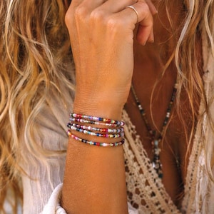 May include: A stack of beaded bracelets in various colors, including red, blue, green, and yellow. The bracelets are worn on a wrist, showcasing the colorful beads and their arrangement. The bracelets are a bohemian style.