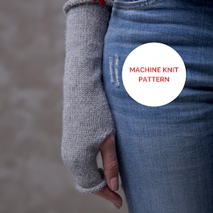 Könnte beinhalten: Ein Paar graue Fingerlose Handschuhe mit einem weißen Kreis, auf dem "MACHINE KNIT PATTERN" steht, auf einer blauen Jeans.