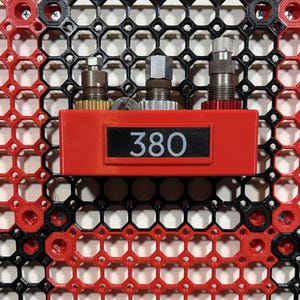 May include: A red plastic component with the number "380" displayed on a black label. Three metal components are mounted above the label. The background is a black and red honeycomb pattern.