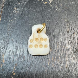 Pode incluir: Um pequeno pingente retangular branco sujo com um anel de latão. O pingente tem uma série de pequenas reentrâncias circulares. O pingente está em uma superfície escura e texturizada.