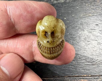 May include: A small, carved skull pendant in shades of tan and brown. The skull has a wide, toothy grin and detailed facial features. The pendant is held between fingers, showcasing its size and intricate design.