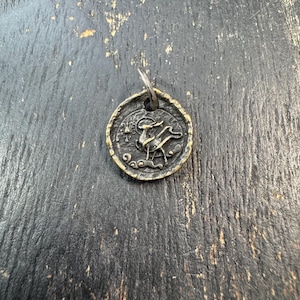 May include: A small, round, antique brass pendant with a detailed relief of a deer-like creature. The pendant has a textured border and a small loop for a chain. The background is a dark, weathered wooden surface.