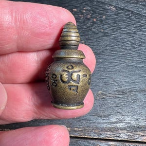 Könnte beinhalten: Ein kleines, antikes bronzefarbenes Gefäß mit rundem Körper und konischem Deckel. Das Gefäß weist geätzte Symbole und Zeichen um seinen Körper auf. Das Objekt wird in einer Hand vor einem dunklen Holzgrund gehalten.