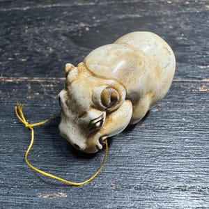 May include: A small, off-white ceramic figurine of a ram with curled horns and a yellow string attached. The ram is lying down on a dark, weathered wooden surface. The figurine has detailed features and a slightly aged appearance.