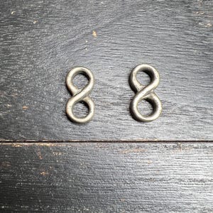 May include: Two silver-coloured infinity loop charms. The charms are made of metal and have a smooth, rounded appearance. They are displayed against a dark, textured wooden surface. These charms could be used for jewellery making or other craft projects.