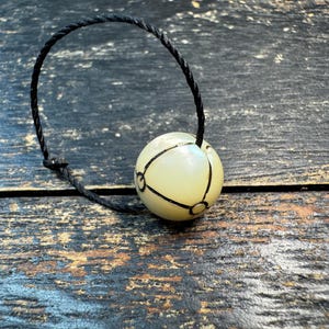 Peut inclure: Une perle sphérique blanc cassé lisse avec un motif de lignes géométriques noires, fixée à un cordon noir torsadé fin. La perle présente une finition brillante et est suspendue par le cordon, qui forme une boucle au-dessus de l'élément sphérique.
