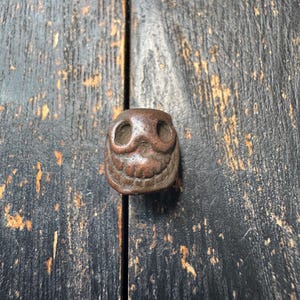 Puede incluir: Pequeño objeto decorativo en forma de búho, de color marrón. El búho tiene dos grandes cuencas oculares y una boca sonriente. El objeto está hecho de un material texturizado y parece ser vintage o antiguo. El fondo es una superficie de madera oscura.