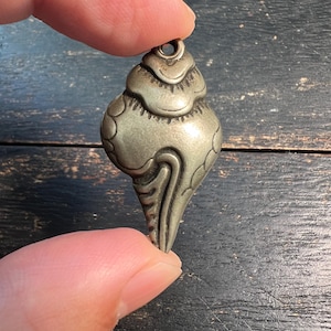 Könnte beinhalten: Ein silberfarbener Anhänger mit einem einzigartigen, stilisierten Design. Der Anhänger hat eine bauchige Oberseite mit strukturierter Oberfläche und eine spitz zulaufende, fließende Unterseite. Er wird von einem Finger vor einem dunklen Holzgrund gehalten.