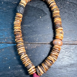 以下が含まれることがあります： 小さくて丸い、自然な色合いのビーズが連なった手作りのネックレス。ビーズは円形に配置され、暗い彫刻ビーズと明るい色の石ビーズがあります。下部には、バーガンディ色のタッセルが付いています。