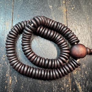 May include: Dark brown wooden prayer beads with a large, round bead and a beige tassel. The beads are circular and strung together, creating a looped design. The background is a dark wooden surface.