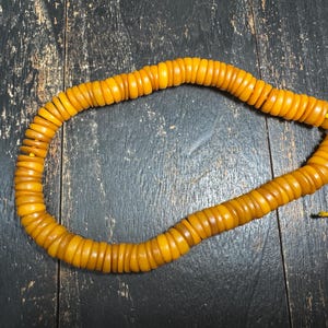 May include: A strand of circular, amber-colored beads strung on a thin yellow cord. The beads are uniform in shape and size, featuring a smooth, polished finish. The strand is arranged in a loose, open loop, showcasing the consistent golden-yellow hue and the simple, functional design of the stringing material.