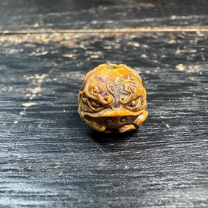 May include: A detailed, carved wooden frog figurine. The frog is a warm, golden brown color with intricate patterns and designs carved into its surface. The frog's eyes are visible, and the overall design is ornate and detailed.