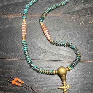 May include: A handcrafted necklace featuring turquoise and brown beads, accented with brass elements. The necklace includes several larger, patterned beads and a decorative brass pendant. The necklace is displayed on a dark wooden surface.