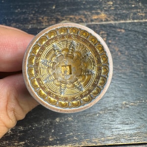 Puede incluir: Un objeto metálico circular con acabado dorado que presenta un intrincado diseño en relieve. El centro muestra un recorte cuadrado rodeado de patrones radiantes y un borde de pequeñas formas redondeadas similares a pétalos.
