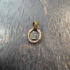 Puede incluir: Un pequeño colgante redondo de color bronce con un lazo dorado. El colgante presenta una figura sentada en posición de meditación. El fondo es una superficie oscura y texturizada.