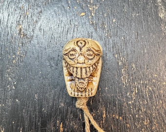 May include: A carved, beige-colored pendant with a stylized face design. The pendant has a detailed, textured surface and is attached to a natural-colored twine cord. The background is a dark, weathered wooden surface.