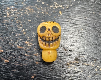 May include: A small, carved, yellowish-tan skull bead with a wide, grinning expression. The eye sockets and mouth are filled with dark, recessed detailing, and the piece features a cylindrical base with a small hole, resembling a decorative charm.