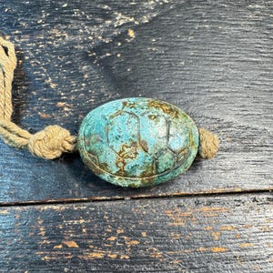 May include: A weathered, turquoise-colored turtle-shaped object with a rope attached. The turtle has a textured surface with a geometric pattern. The rope is a natural beige color and knotted at the end. The object is on a dark wooden surface.