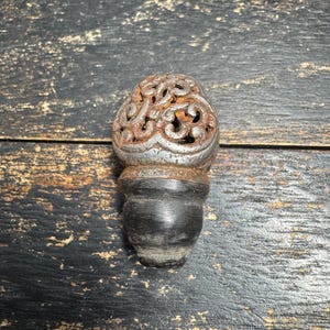 Könnte beinhalten: Ein dekorativer Endknopf mit einem abgerundeten, durchbrochenen Metalloberteil in rostiger, silberfarbener Optik mit komplexen Schnörkelmustern. Der Sockel besteht aus einem glatten, dunklen, massiven Material mit abgerundeter, konischer Form.